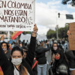 Featured image: Protester in Colombia holding a banner that reads: "In Colombia they are killing us, Uribe gave the order." Photo courtesy of RedRadioVe.