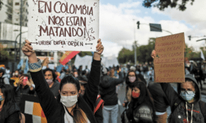 Featured image: Protester in Colombia holding a banner that reads: "In Colombia they are killing us, Uribe gave the order." Photo courtesy of RedRadioVe.