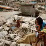 Haitian woman with a baby in her lap seating over earthquake rubble. Photo by Twitter /@KhaledBeydoun .