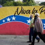 Two elder Venezuelans wearing face masks walk in front of a mural with the face of Simon Bolivar, the Venezuelan flag and with a "long live the homeland" caption. Photo by EFE.