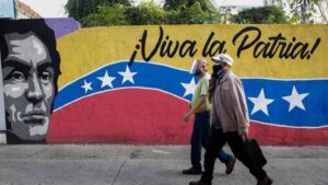 Two elder Venezuelans wearing face masks walk in front of a mural with the face of Simon Bolivar, the Venezuelan flag and with a "long live the homeland" caption. Photo by EFE.