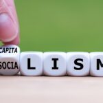 Hand flips a dice and changes the word “Socialism” to “Capitalism”, or vice versa. (Fokusiert / Getty Images).