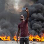 Protester in Haiti taking a selfie with burning fires in the background. File photo by Odelyn Joseph/AP/DPA.
