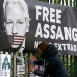 Activist adding placards near a big banner with a photo of Julian Assange with his mouth strapped with the US flag. The banner reads "Free Assange, No Extradition." Photo: Reuters/Henry Nicholls.