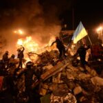 Featured image: State flag of Ukraine behind a wall of anonymous protesters in Kyiv, Ukraine (February 18, 2014). By Mstyslav Chernov/Unframe – Own work, CC BY-SA 3.0, Link.