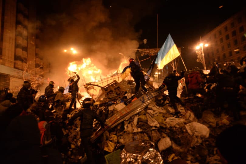 Featured image: State flag of Ukraine behind a wall of anonymous protesters in Kyiv, Ukraine (February 18, 2014). By Mstyslav Chernov/Unframe – Own work, CC BY-SA 3.0, Link.