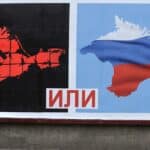 Passerby looking at a billboard with two maps of Ukraine, one to the left marked with a svastica and another to the right with the Russian flag, divided by the Russian text “или” between the two maps meaning “or”. Photo: PBS.