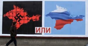 Passerby looking at a billboard with two maps of Ukraine, one to the left marked with a svastica and another to the right with the Russian flag, divided by the Russian text “или” between the two maps meaning “or”. Photo: PBS.