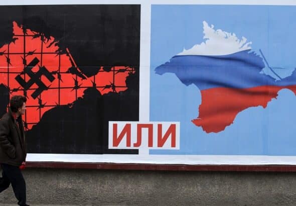 Passerby looking at a billboard with two maps of Ukraine, one to the left marked with a svastica and another to the right with the Russian flag, divided by the Russian text “или” between the two maps meaning “or”. Photo: PBS.