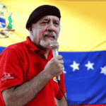 Carlos Lanz giving a speech wearing a red shirt and black beret, with a Venezuelan flag in the background. File photo.