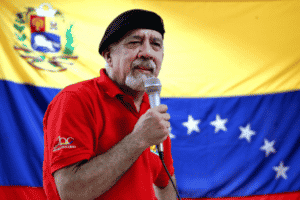 Carlos Lanz giving a speech wearing a red shirt and black beret, with a Venezuelan flag in the background. File photo.