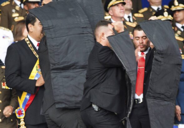 In this photo released by China's Xinhua News Agency, security personnel surround Venezuela's President Nicolás Maduro, left, after drones armed with explosives detonated as he was making a speech in Caracas, Venezuela, Aug. 4, 2018.