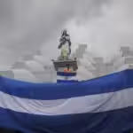 Giant Nicaraguan flag waving below the figure of a saint and a person holding a small Nicaraguan flag. File photo.