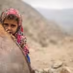 Yemeni girl with an atribulated look shows her face will covering behind a metal object, Yemeni mountains in the background. Photo: Kellie Ryan/IRC.
