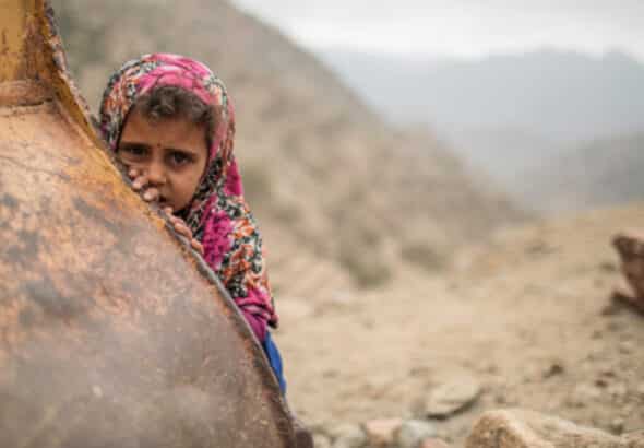 Yemeni girl with an atribulated look shows her face will covering behind a metal object, Yemeni mountains in the background. Photo: Kellie Ryan/IRC.