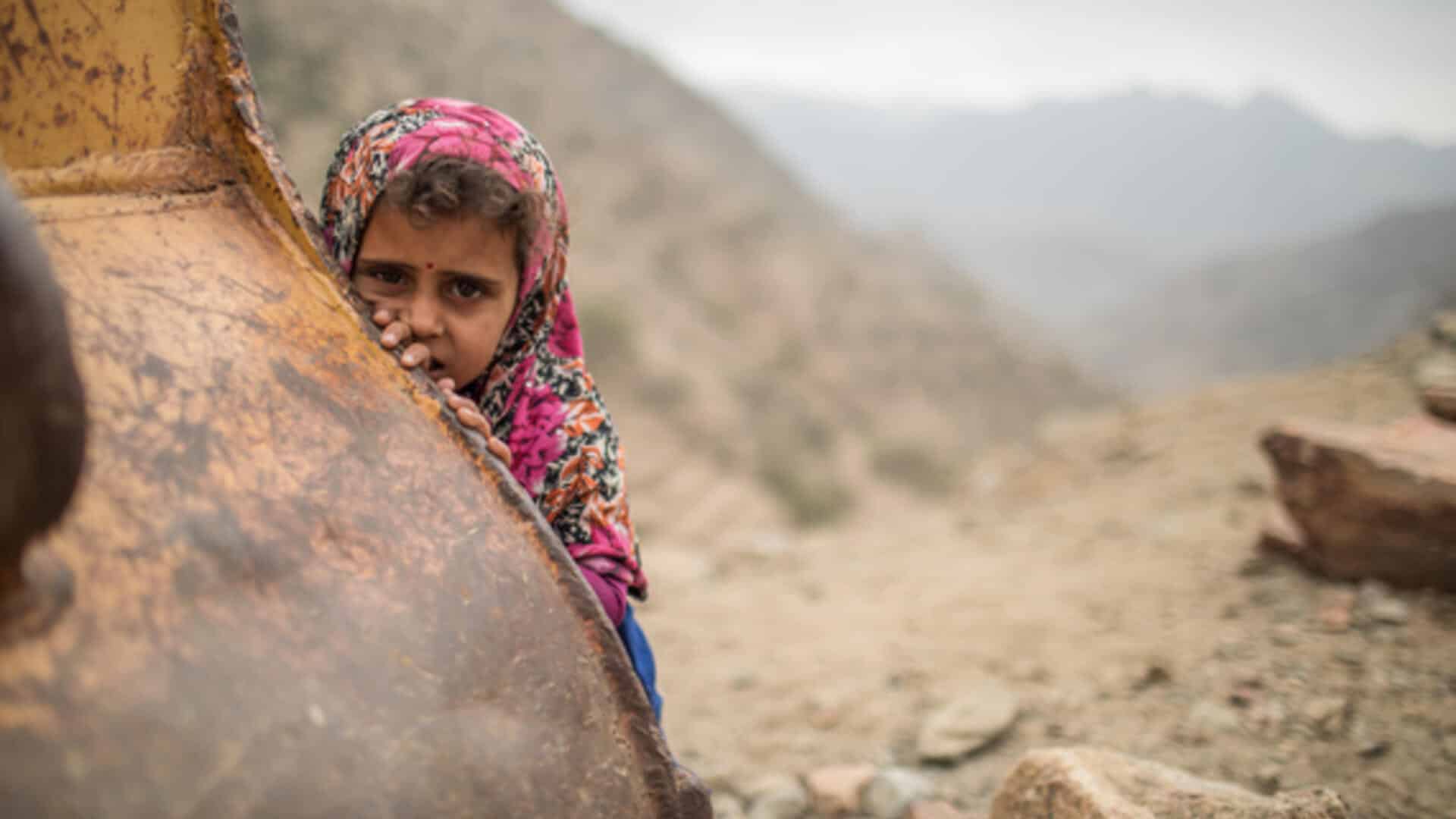 Yemeni girl with an atribulated look shows her face will covering behind a metal object, Yemeni mountains in the background. Photo: Kellie Ryan/IRC.