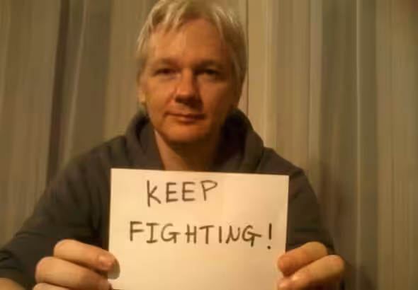Julian Assange with a poster that reads "keep fighting." Photo: Peoples Dispatch.