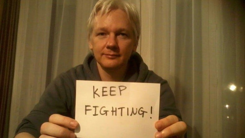 Julian Assange with a poster that reads "keep fighting." Photo: Peoples Dispatch.