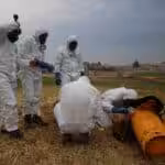 People with biohazard suits making an inspection of a orange object in a field. Photo: OPCW.