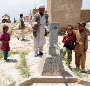 Afghan man surrounded by kids pushing a water pump but no water seen from the other side. A US military truck can be seen in the background. Photo: Kelley J. Stewart via ISAF Headquarters Public Affairs Office from Kabul, Afghanistan – CC BY 2.0.