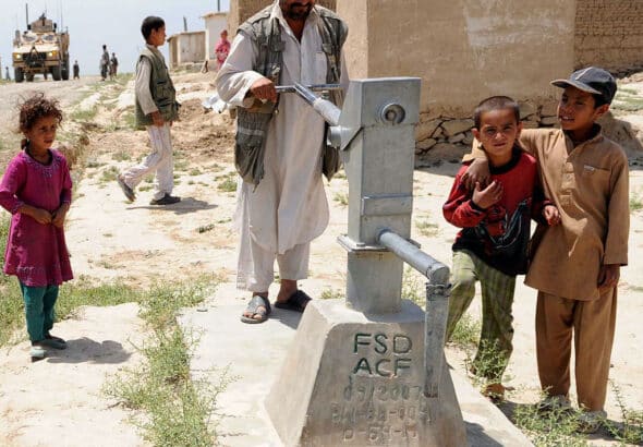 Afghan man surrounded by kids pushing a water pump but no water seen from the other side. A US military truck can be seen in the background. Photo: Kelley J. Stewart via ISAF Headquarters Public Affairs Office from Kabul, Afghanistan – CC BY 2.0.