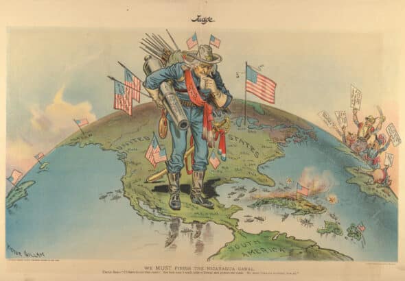 Early 20th century cartoon depicting an “Uncle Sam” carrying a cannon and multiple US flags, stepping over Texas in a map of the hemisphere, and looking down to Central and South America in a reflexive way. Cartoon by Victor Guillam/Cornell University.