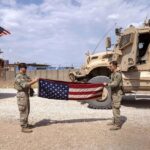 Two US soldiers folding their flag and in the back two war trucks. Photo: The Cradle.