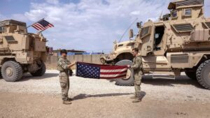 Two US soldiers folding their flag and in the back two war trucks. Photo: The Cradle.