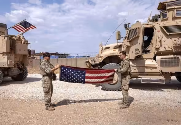 Two US soldiers folding their flag and in the back two war trucks. Photo: The Cradle.
