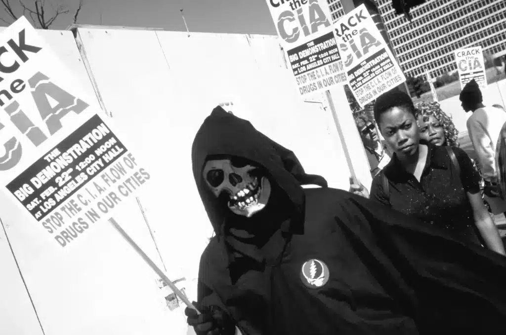 People protesting and holding signs, one of them dressed in a dead costume and holding a sign that reads: "CIA - The big demonstration, Sat. Feb. 22nd at 12:00 noon at Los Angeles City Hall - Stop the flow of drugs from the CIA in our cities." Photo: Rainer's Newsletter.