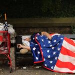 A homeless person in the United States sleeping on a park bench, wrapped in a blanket decorated with the pattern of the US flag. Photo: Spencer Platt/Getty Images.