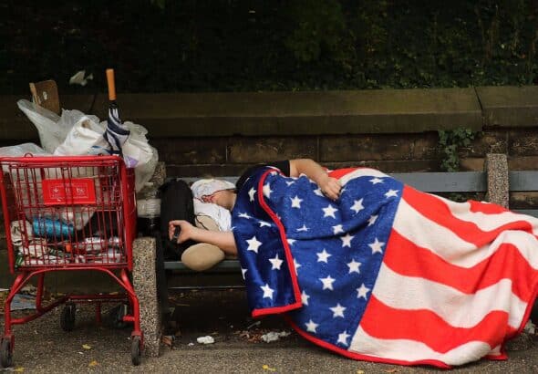 A homeless person in the United States sleeping on a park bench, wrapped in a blanket decorated with the pattern of the US flag. Photo: Spencer Platt/Getty Images.
