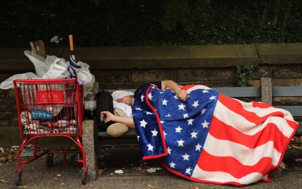 A homeless person in the United States sleeping on a park bench, wrapped in a blanket decorated with the pattern of the US flag. Photo: Spencer Platt/Getty Images.