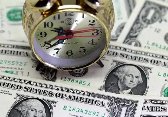 An analog alarm clock sits on top of a spread of one dollar bills. Photo: CGTN.