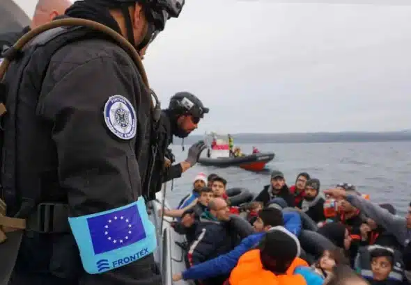 A FRONTEX official stands before a boat carrying migrants. Photo: MasarBadil.org.