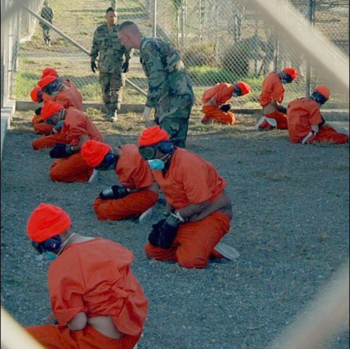 Detainees in orange jumpsuits sit in a holding area under the "watchful eyes" of Military Police at Camp X-Ray at Naval Base Guantanamo Bay, Cuba, during in-processing to the "temporary detention facility" on Jan. 11, 2002. Photo: Shane T. McCoy, U.S. Navy via Wikimedia Commons.