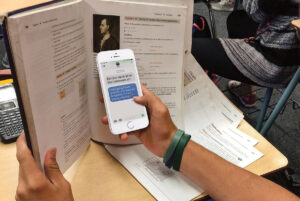 Student in class texting on a cellphone that is hidden by a larger book. Photo: Scholastic/file photo.