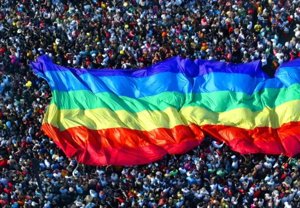 An LGBTQIA+ Pride flag is carried by a marching crowd of supporters. Photo: Getty Images.