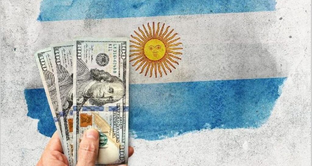 Three American 100 dollar bills with a blurred Argentinian flag in the background. Photo: El Cronista/File photo.