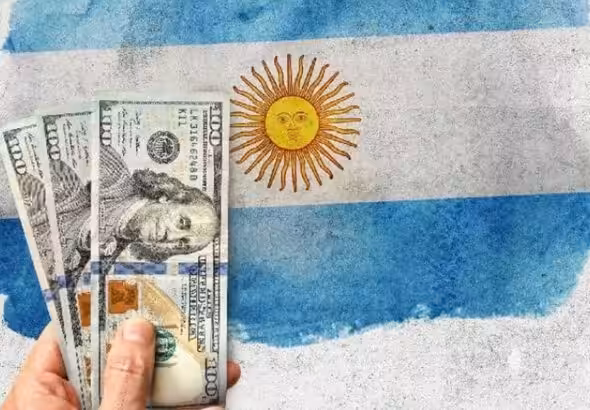 Three American 100 dollar bills with a blurred Argentinian flag in the background. Photo: El Cronista/File photo.
