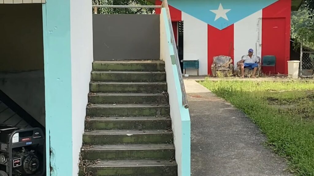 Outdoor stairs in a house and a man sitting in a worn-out armchairs in the background with the Puerto Rican flag painted on a wall. Photo: Geopolitical economy/File photo.