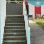 Outdoor stairs in a house and a man sitting in a worn-out armchairs in the background with the Puerto Rican flag painted on a wall. Photo: Geopolitical economy/File photo.