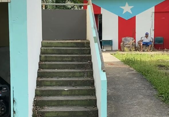 Outdoor stairs in a house and a man sitting in a worn-out armchairs in the background with the Puerto Rican flag painted on a wall. Photo: Geopolitical economy/File photo.