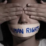 Young woman with her eyes covered with her hands and her mouth strapped with adhesive tape with a caption that reads Human Rights. Photo: Caitlin Johnstone/File Photo.