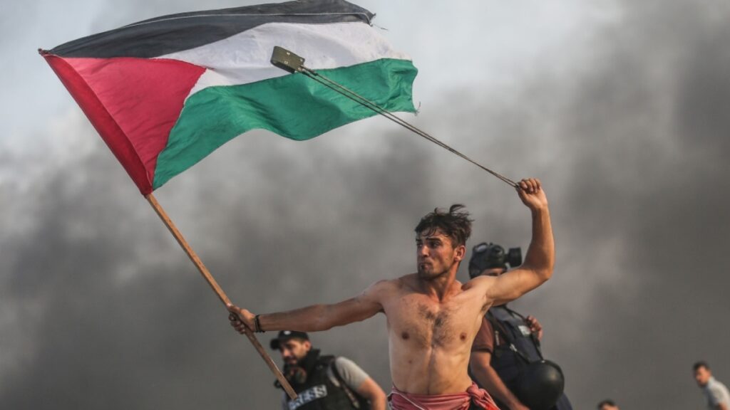 A man waves the Palestinian flag and throws rocks with a probe. Photo: Mustafa Hassouna.