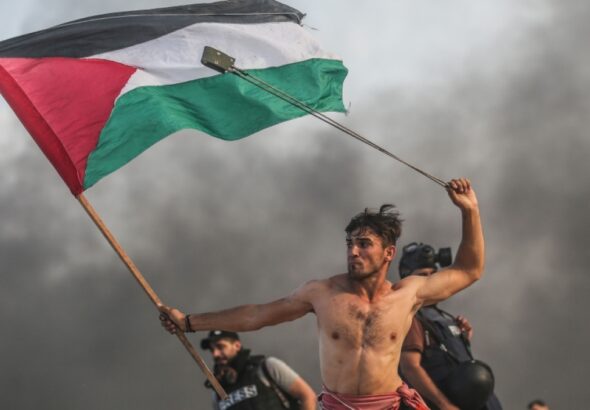 A man waves the Palestinian flag and throws rocks with a probe. Photo: Mustafa Hassouna.