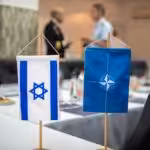 A NATO and an Israeli table side flags on a blurred buffet table and two military officers talking. Photo: NATO Maritime Command/File photo.