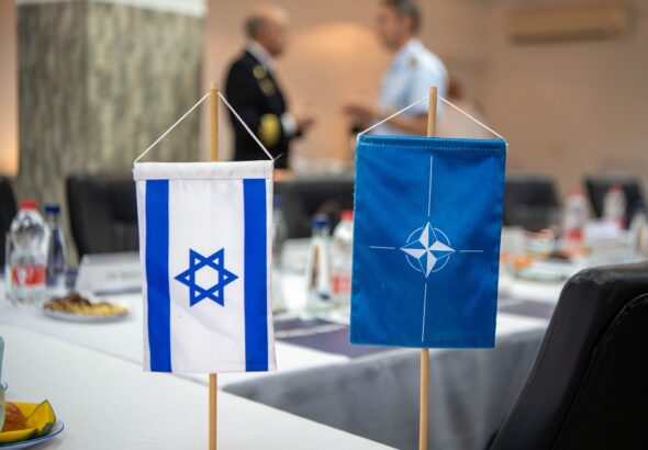 A NATO and an Israeli table side flags on a blurred buffet table and two military officers talking. Photo: NATO Maritime Command/File photo.