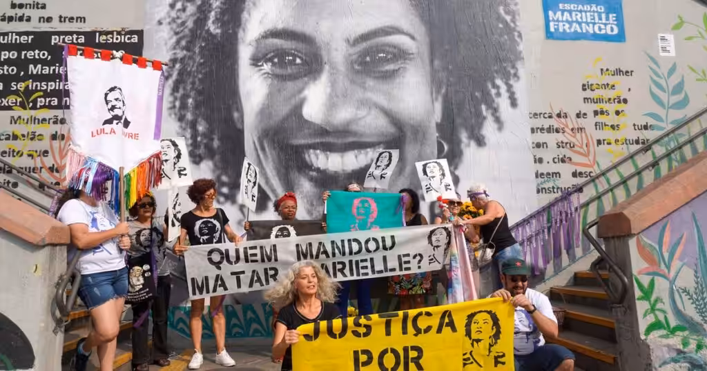 People holding banners and posters demanding justice for Marielle Franco and asking who is behind her assassination, in front of a mural photo of the Brazilian LGBTQ activist and politician. Photo: Scuzinska/Alamy Stock Photo/File photo.