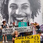 People holding banners and posters demanding justice for Marielle Franco and asking who is behind her assassination, in front of a mural photo of the Brazilian LGBTQ activist and politician. Photo: Scuzinska/Alamy Stock Photo/File photo.